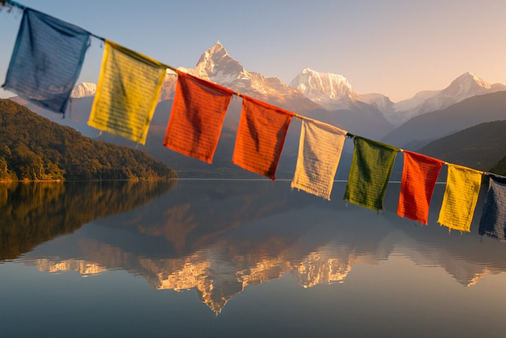 Die schneebedeckten Himalaya-Gipfel des Annapurna-Massivs spiegeln sich bei Sonnenaufgang im Phewa-See bei Pokhara, Nepal, mit bunten Gebetsfahnen